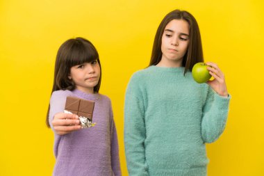 Sarı arka planda izole edilmiş küçük kız kardeşler bir elinde çikolata tableti, diğerinde elma alıyorlar.