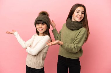 Pembe arka planda izole edilmiş kışlık giysiler giyen küçük kız kardeşler davet edilmek için ellerini yana uzatıyor.