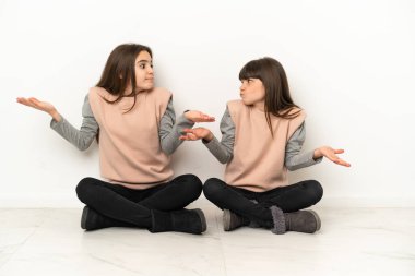 Beyaz arka planda izole bir şekilde yerde oturan küçük kız kardeşler omuzlarını kaldırırken önemsiz jestler yapıyorlar.