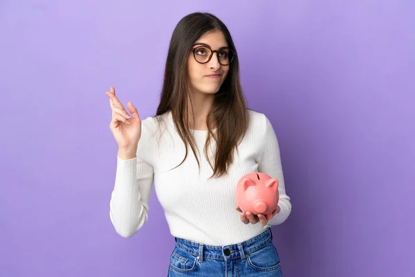 Young Caucasian Woman Holding Piggybank Isolated Purple Background Fingers Crossing — Stock Photo, Image