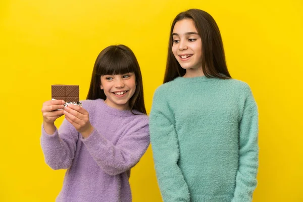 Sarı arka planda izole edilmiş küçük kız kardeşler çikolata tableti alıyor ve mutlular.