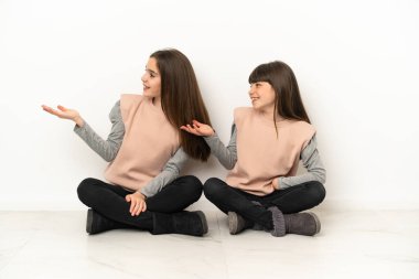 Beyaz arka planda izole bir şekilde yerde oturan küçük kardeşler geriye dönük göstererek bir ürün sunuyorlar.