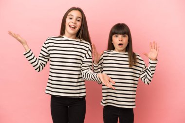 Küçük kız kardeşler pembe arka planda izole edilmiş, şaşırmış ve şok olmuş yüz ifadeleriyle.
