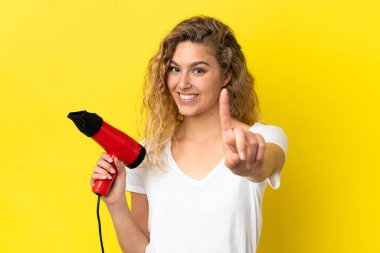 Sarı arka planda izole edilmiş saç kurutma makinesi tutan genç sarışın kadın parmağını kaldırıyor.