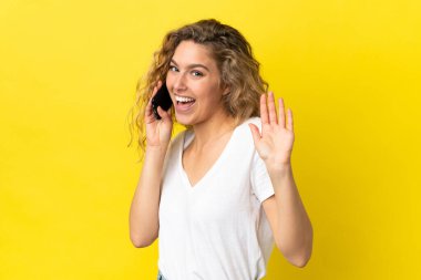 Sarı arka planda izole edilmiş cep telefonu kullanan genç sarışın kadın mutlu bir ifadeyle selamlıyor.