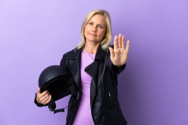 Mor arka planda izole edilmiş motosiklet kaskı tutan orta yaşlı bir kadın dur hareketi yapıyor.