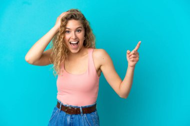 Mavi arka planda izole edilmiş genç sarışın kadın şaşırmış ve işaret ediyor.