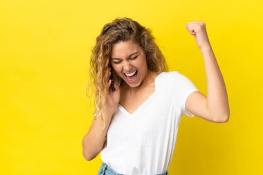 Sarı arka planda cep telefonu kullanan genç sarışın kadın zaferi kutluyor.