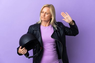 Mor arka planda motosiklet kaskı tutan orta yaşlı bir kadın dur hareketi yapıyor ve hayal kırıklığına uğruyor.