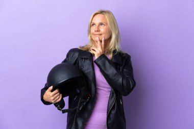 Mor arka planda izole edilmiş motosiklet kaskı tutan orta yaşlı bir kadın gülümserken yukarı bakıyor.