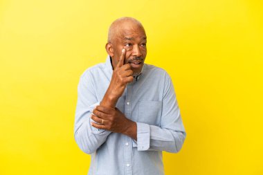 Küba Senior sarı arka planda izole edilmiş bir şekilde bir ürün sunmak ve bir şeyler fısıldamak için kenarı işaret ediyor.