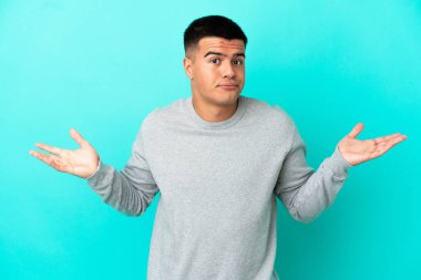 Soyutlanmış mavi arka planı olan genç yakışıklı adam ellerini kaldırırken şüphe duyuyor.