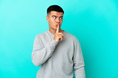 Soyutlanmış mavi arka planda genç yakışıklı bir adam sessizlik işareti gösteriyor. Parmağını ağzına sok.