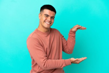 İzole edilmiş mavi arka plan üzerinde genç yakışıklı bir adam bir ilan vermek için kopyalama alanı tutuyor