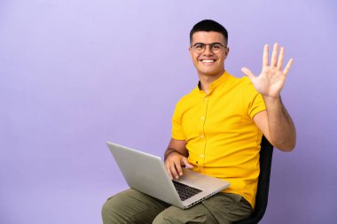 Dizüstü bilgisayarlı bir sandalyede oturan genç adam parmaklarıyla beşi sayıyor.