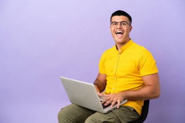 Genç adam dizüstü bilgisayarlı bir sandalyede oturuyor ve yüzünde sürpriz bir ifade var.