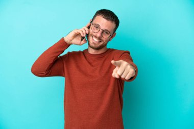 Mavi arka planda izole edilmiş cep telefonu kullanan Brezilyalı genç adam mutlu bir ifadeyle öne işaret ediyor.