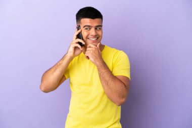 İzole edilmiş mor arka planda cep telefonu kullanan yakışıklı genç adam yana bakıyor ve gülümsüyor.