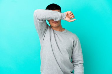 İzole edilmiş mavi arka planda genç yakışıklı adam gözlerini elleriyle kapatıyor.