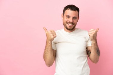 Pembe arka planda izole edilmiş genç Brezilyalı adam başparmakları havada ve gülümsüyor.