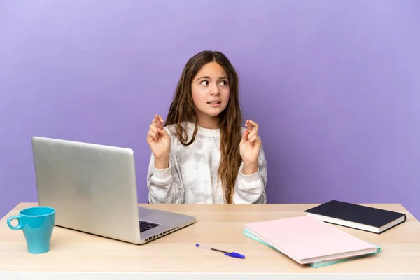 Little student girl in a workplace with a laptop isolated on purple background with fingers crossing and wishing the best
