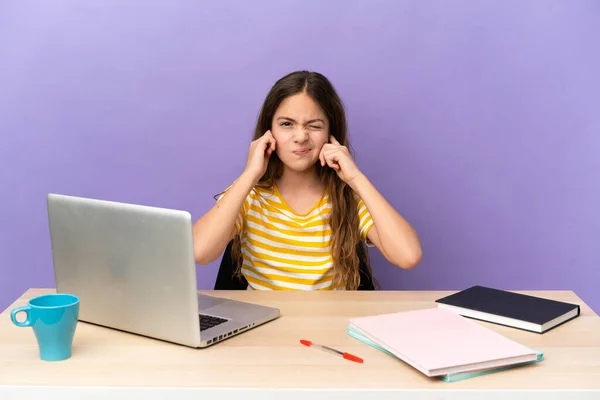 Little student girl in a workplace with a laptop isolated on purple background frustrated and covering ears