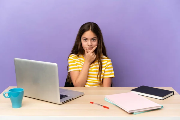 Little student girl in a workplace with a laptop isolated on purple background thinking