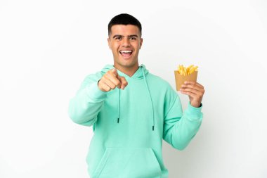 Genç yakışıklı adam izole edilmiş beyaz arka planda patates kızartması tutuyor şaşırmış ve ön tarafı işaret ediyor.