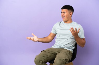 İzole edilmiş arka planda oturan genç bir adam. Sürpriz bir yüz ifadesi var.