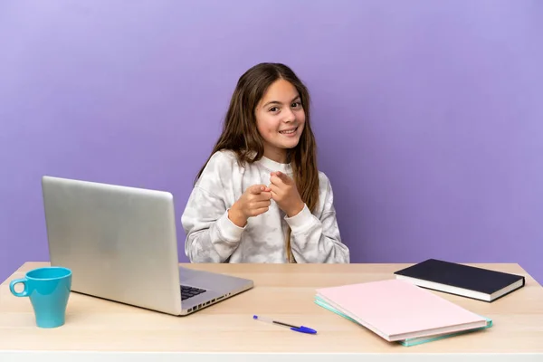 Little student girl in a workplace with a laptop isolated on purple background surprised and pointing front