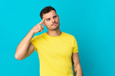 Genç, beyaz, yakışıklı bir adam mavi arka planda izole edilmiş. Delilik jesti yapıyor. Parmağını kafaya koyuyor.