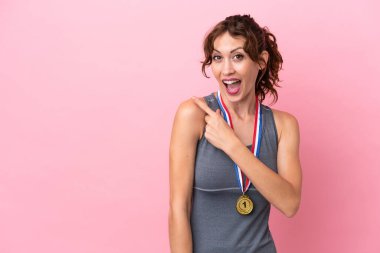 Pembe arka planda madalyaları olan genç beyaz kadın şaşırmış ve işaret ediyor.