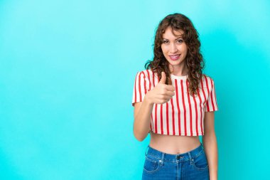 Mavi arka planda, kıvırcık saçlı genç bir kadın baş parmak hareketi yapıyor.