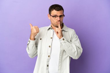 İzole edilmiş mor arka planı olan Brezilyalı adam yan tarafı işaret ediyor ve sessizlik hareketi yapıyor.