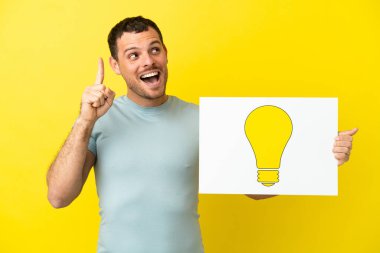 İzole edilmiş mor arka planı olan Brezilyalı bir adam elinde ampul simgesi olan bir levha tutuyor ve düşünüyor.