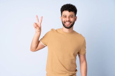 Mavi arka planda izole edilmiş genç Faslı adam gülümsüyor ve zafer işareti gösteriyor.