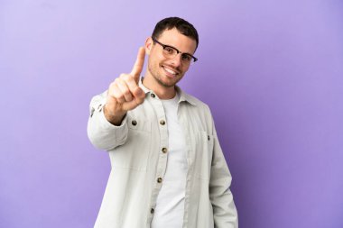 İzole edilmiş mor arka planı olan Brezilyalı adam parmağını kaldırıyor ve gösteriyor.