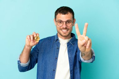 Brezilya 'lı bir adam mavi arka planda Bitcoin tutuyor gülümsüyor ve zafer işareti gösteriyor