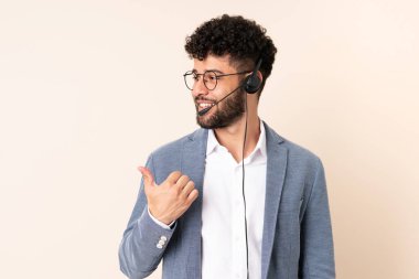 Faslı bir pazarlamacı bej arka planda izole edilmiş bir kulaklıkla çalışıyor. Ürünü sunmak için yan tarafı işaret ediyor.