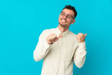 Mavi arka planda izole edilmiş beyaz, yakışıklı genç bir adam bir ürün sunmak için kenarı işaret ediyor.