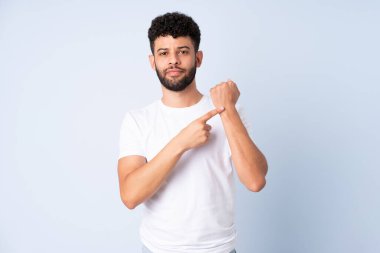 Faslı genç bir adam mavi arka planda izole edilmiş. Geç kalma jesti yapıyor.