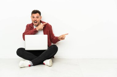 Genç, yakışıklı, beyaz bir adam dizüstü bilgisayarla yerde oturuyor. Şaşırmış ve işaret ediyor.