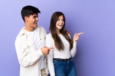 İzole edilmiş mor arka planda genç bir çift, parmak kenarı işaret ediyor.