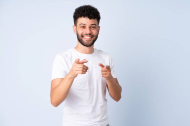 Faslı genç bir adam mavi arka planda izole edilmiş şaşırmış ve ön tarafı işaret ediyor.