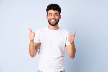 Genç Faslı bir adam mavi arka planda izole edilmiş. Baş parmakları havada ve gülümsüyor.