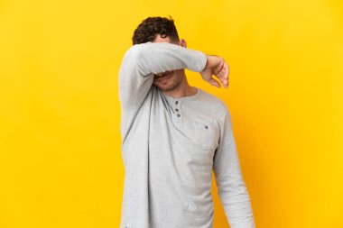 Beyaz tenli yakışıklı adam sarı arka planda izole edilmiş gözlerini elleriyle kapatıyor.