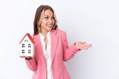 Rus emlakçı, beyaz arka planda bir oyuncak evi tutuyor. Sürpriz bir ifadeyle yan yana bakarken.