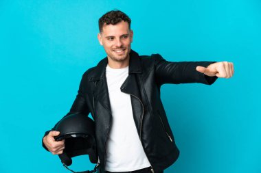 Motosiklet kasklı beyaz bir adam mavi arka planda izole edilmiş bir şekilde baş parmak işareti yapıyor.