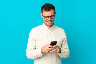 Mavi arka planda izole genç beyaz adam cep telefonuyla bir mesaj gönderiyor.