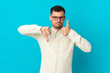 Beyaz tenli yakışıklı adam mavi arka planda izole edilmiş, iyiye-kötüye işaret yapıyor. Evet ya da hayır arasında kararsız